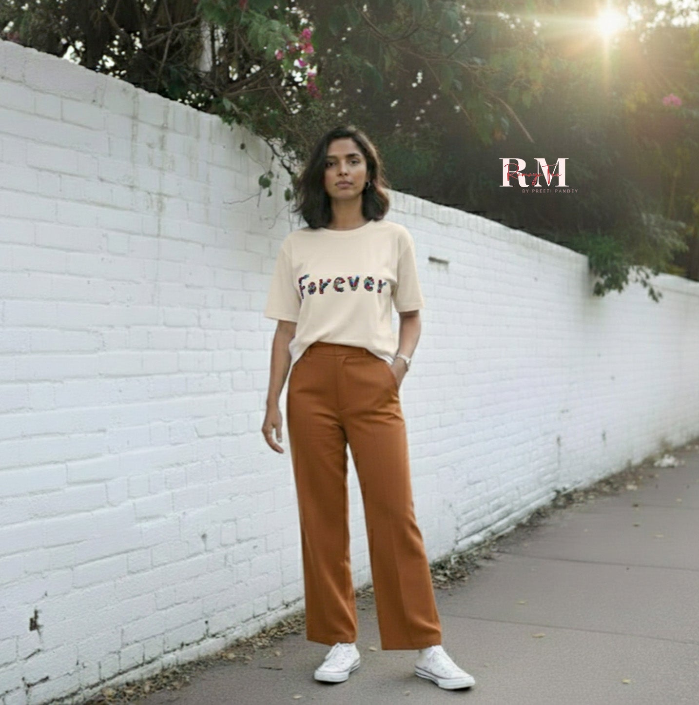 Woman wearing a beige sweater with text and brown pants standing against a white brick wall.