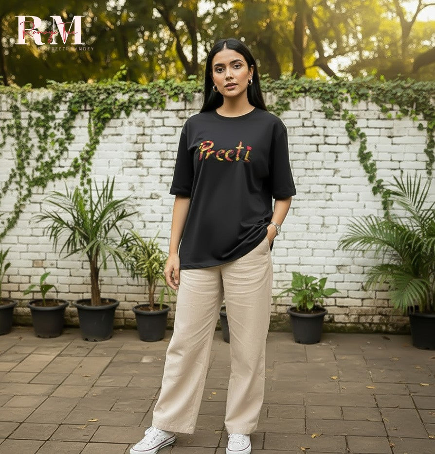 Person wearing a black t-shirt with colorful text, standing outdoors against a white brick wall with greenery.