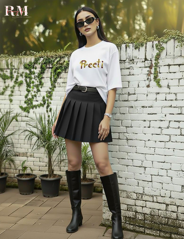 Woman wearing a white top with 'Preeti' printed on it, black pleated skirt, and black knee-high boots standing against a white brick wall.