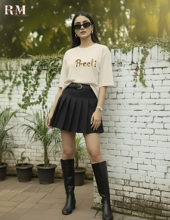 Woman wearing a beige top with text and black pleated skirt, standing in front of a white brick wall with plants.