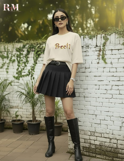 Woman wearing a beige top with text and black pleated skirt, standing in front of a white brick wall with plants.