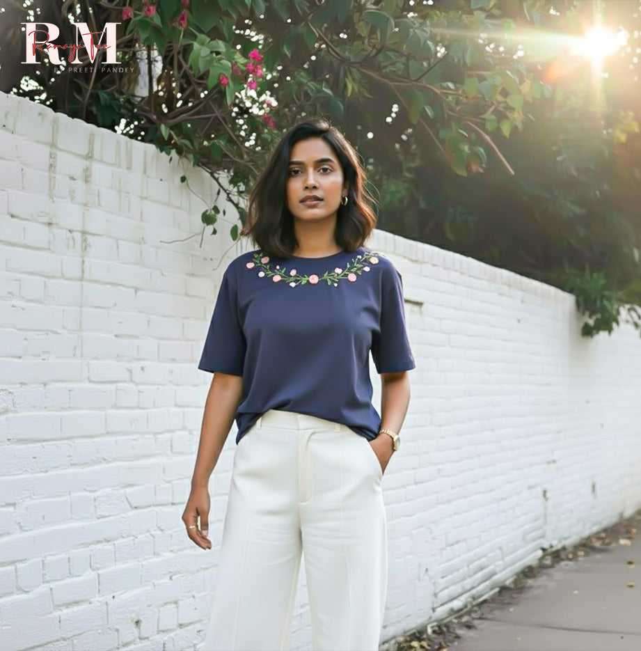 Woman wearing a navy blue top with floral embellishments and white pants standing against a white brick wall.