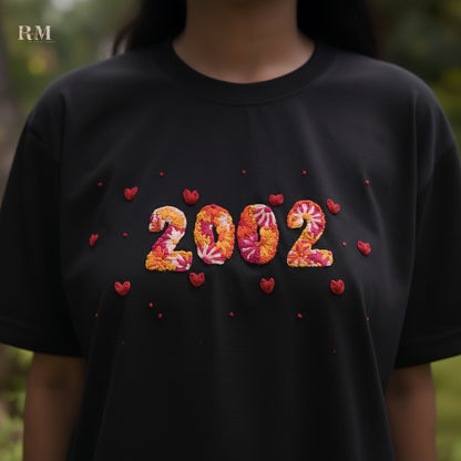 Black t-shirt with '2002' design featuring flowers and hearts on a blurred natural background.