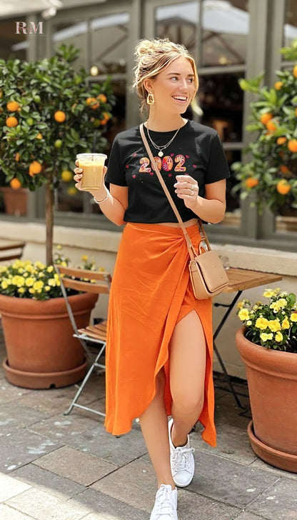 Woman in black t-shirt with '2022' text, orange skirt, and white sneakers holding a drink outdoors.