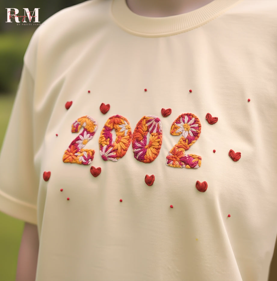 Beige t-shirt with floral '2002' design and red hearts on a blurred green background